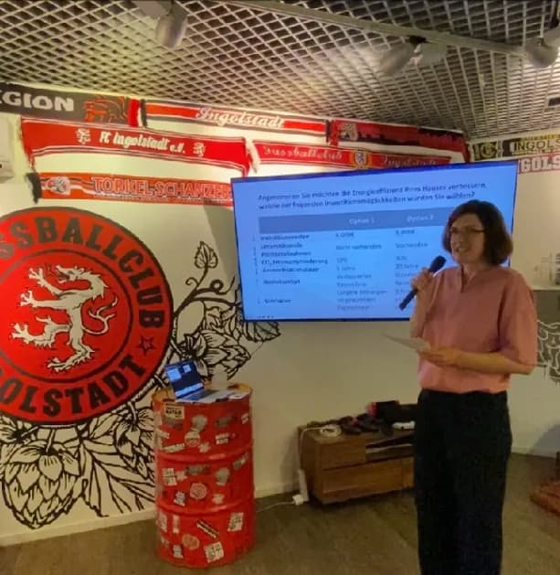 Eine Frau in einem rosa Hemd und einer dunklen Hose steht vor einer Leinwand mit einer Tabelle. Die Tabelle vergleicht zwei Optionen, vermutlich für Investitionen, und listet Kriterien wie Investitionskosten, Umsatzrendite, CO2-Emissionsminderung und Amortisationsdauer auf. Sie hält ein Mikrofon in der Hand und scheint eine Präsentation zu halten. Im Hintergrund sieht man verschiedene Schals und Dekorationen mit dem Logo des Fußballvereins Ingolstadt, was darauf hindeutet, dass die Präsentation in einem Veranstaltungsort stattfindet, der mit diesem Verein in Verbindung steht.