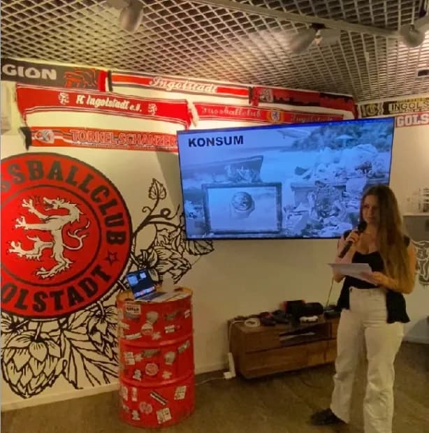 Eine junge Frau steht vor einer Wand mit einem Fußballclub-Logo und einem Bildschirm, auf dem eine Präsentation mit dem Titel "Konsum" läuft. Sie hält ein Mikrofon und Papiere in der Hand, während sie eine Präsentation hält. Ein Laptop steht auf einem roten Fass mit Aufklebern, das als Tisch dient. An der Wand hängen Fußballschals, die das Ambiente des Veranstaltungsortes unterstreichen.