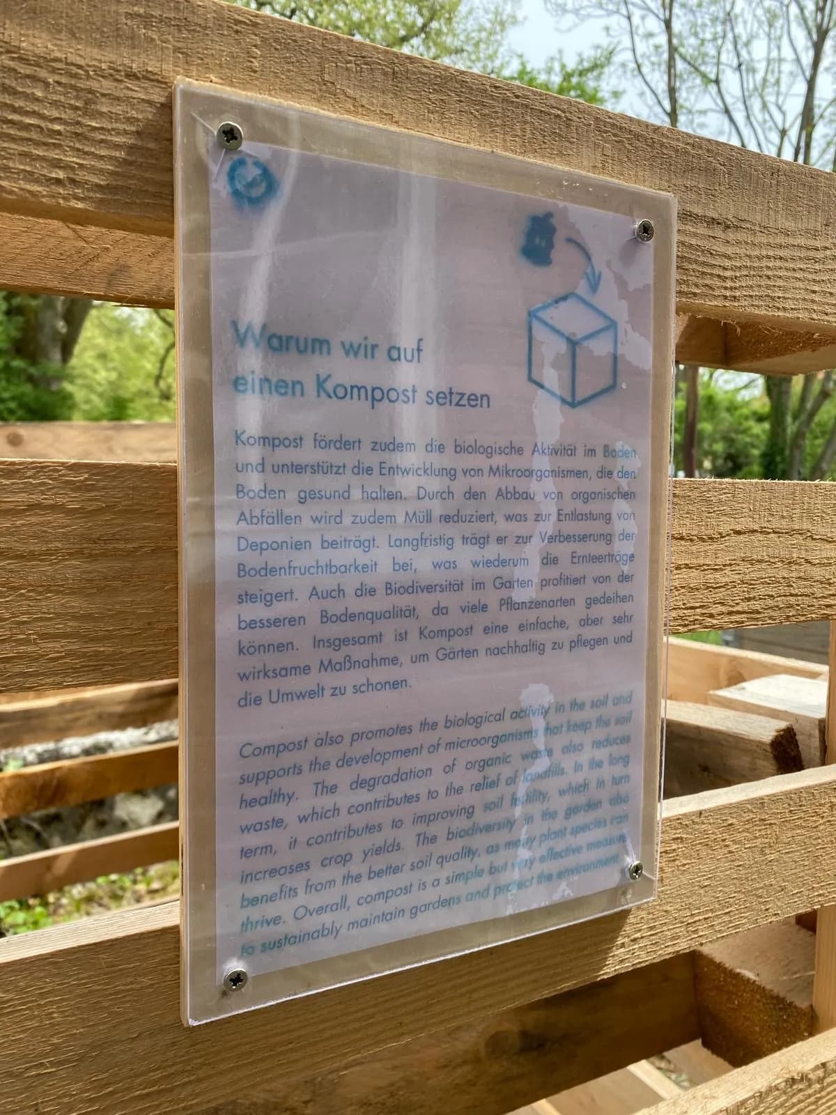 Ein Schild auf einem Holzzaun erklärt die Vorteile des Kompostierens auf Deutsch, einschließlich der Förderung der biologischen Aktivität im Boden, der Unterstützung des Wachstums von Mikroorganismen, der Reduzierung von Müll und der Verbesserung der Bodenfruchtbarkeit.