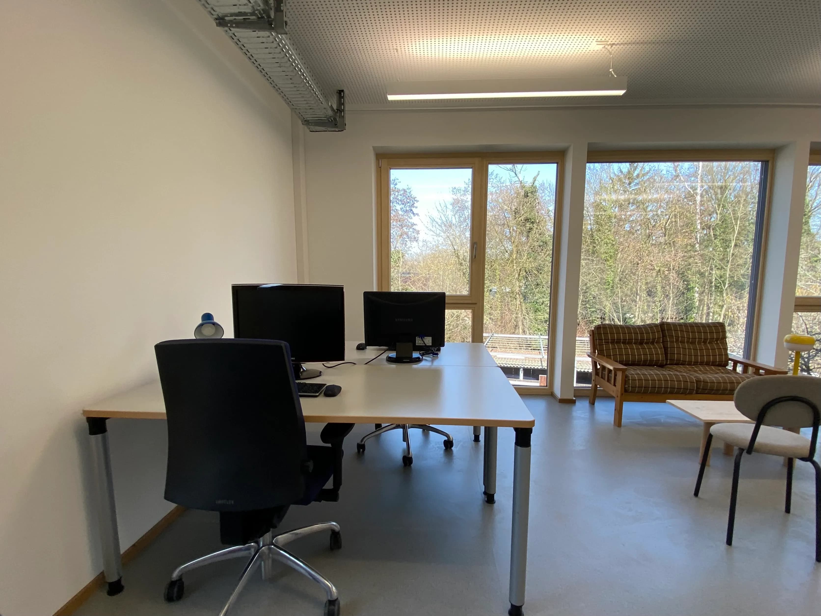 Ein hell erleuchteter, moderner Büroraum mit einem weißen Doppelschreibtisch, zwei Computermonitoren und schwarzen Bürostühlen. Im Hintergrund befindet sich eine Sitzecke mit einem braun karierten Sofa und einem kleinen Tisch vor großen, bodentiefen Fenstern mit Blick auf üppige grüne Bäume.