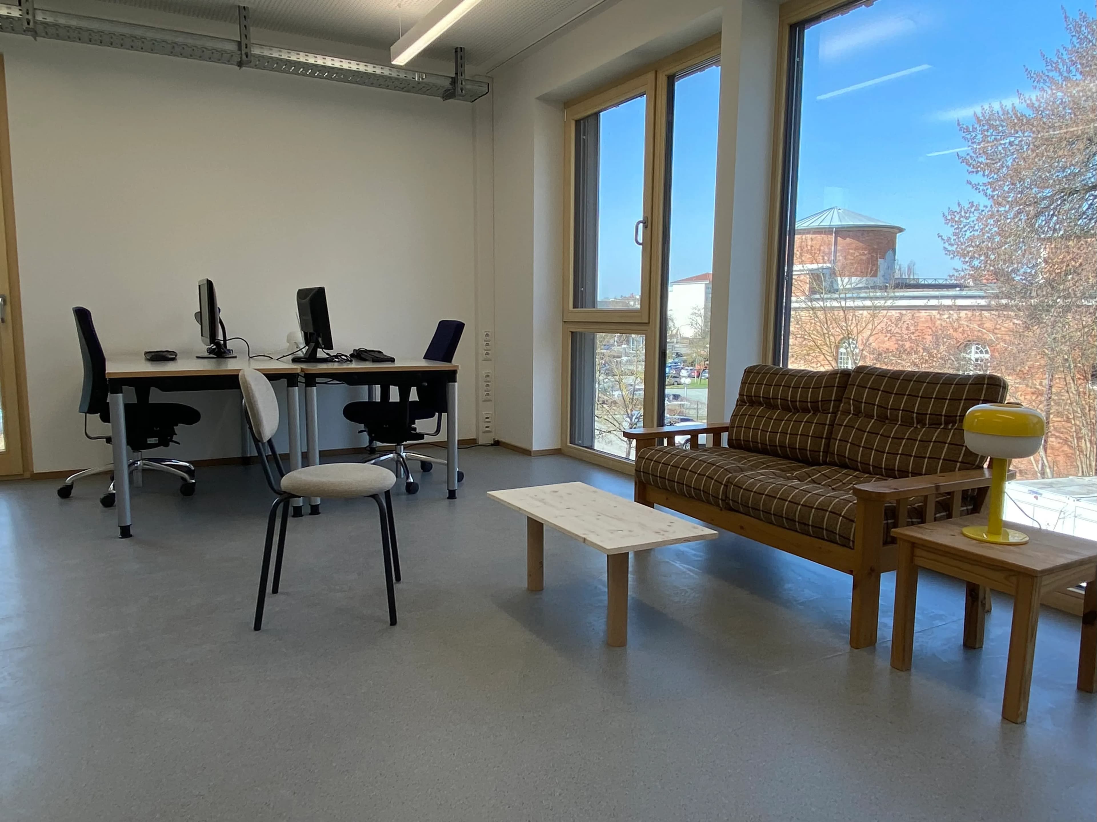 Ein heller, moderner Büroraum mit zwei Schreibtischen und Computermonitoren auf der linken Seite und einem Loungebereich auf der rechten Seite. Die Lounge besteht aus einem braun karierten Sofa, einem minimalistischen Couchtisch aus hellem Holz und einem kleinen Beistelltisch mit einer gelben Lampe. Große, bodentiefe Fenster fluten den Raum mit natürlichem Licht und bieten einen Ausblick auf einen klaren blauen Himmel, blühende Bäume und ein Backsteingebäude mit einem Rundturm in der Ferne.