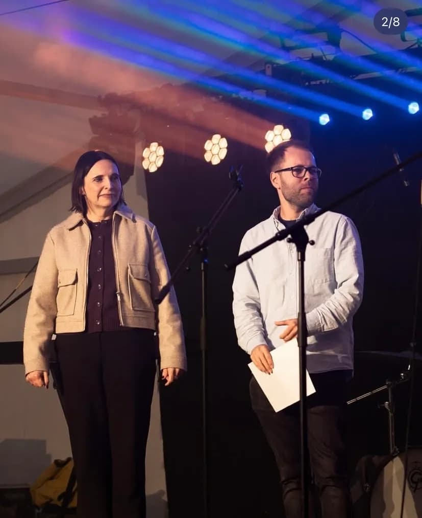 Eine Frau und ein Mann stehen auf einer Bühne unter hellem Licht. Die Frau links trägt eine beige Jacke und eine dunkle Hose, während der Mann rechts eine Brille und ein hellblaues Hemd trägt und ein weißes Blatt Papier hält. Im Hintergrund sind blaue Lichtstrahlen und Bühnenscheinwerfer zu sehen, im Vordergrund stehen Mikrofonständer.